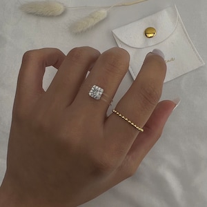 May include: A hand wearing two gold rings. The ring on the index finger has a square-shaped clear stone with smaller stones surrounding it. The ring on the middle finger is a thin band with small gold beads.