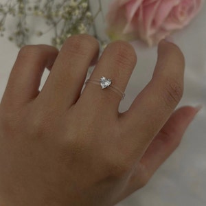 May include: A clear ring with a heart-shaped clear stone. The ring is on a person's finger.