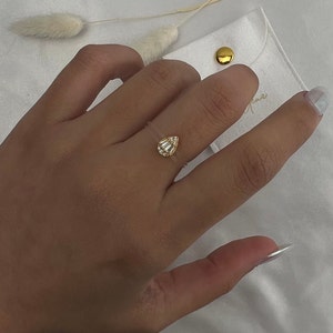May include: A gold ring with a clear teardrop-shaped stone set in the center. The ring is on a finger.