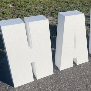 May include: Large, white foam letters spelling the word "HAIR" are displayed outdoors on a paved surface with grass in the background. The letters are thick and blocky, casting shadows.