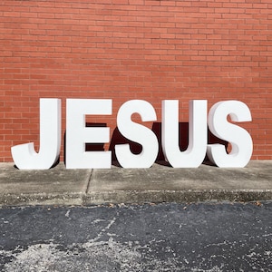May include: White foam letters spelling out the word "JESUS" against a red brick wall background.