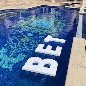 May include: A luxurious swimming pool with a mosaic tile design in shades of blue and turquoise. Large white letters spell out the word "BET" in the water. The pool has a decorative border and is surrounded by a stone patio.