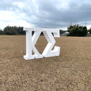 May include: Large white foam letters, spelling out the Greek letters Kappa and Sigma, stand on a field of dry grass. The letters are thick and three-dimensional, with a simple, clean design. The background includes trees and a cloudy sky.