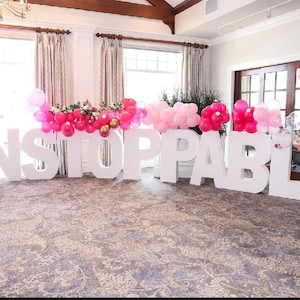 May include: White letters spelling out the word "UNSTOPPABLE" are arranged on a floor. The letters are large and stand upright. Pink and white balloons are strung above the letters.