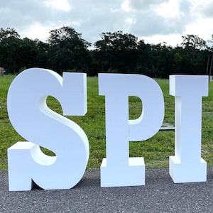 May include: White foam letters spelling out "SPI" in a grassy field. The letters are large and stand upright.