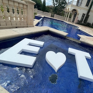 May include: White foam letters "E", a heart, and "T" float in a blue tiled pool. The pool is surrounded by a stone patio and a decorative balustrade. The scene is outdoors with a building in the background.