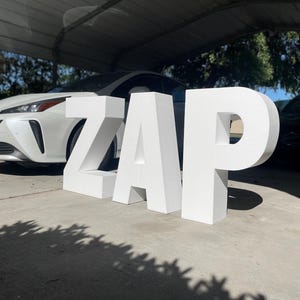 May include: Large, white 3D letters spelling "ZAP" are positioned on a concrete surface. A white car is parked behind the letters, and a black car is partially visible on the right. The scene is outdoors, under a carport.