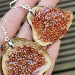 May include: A pair of earrings featuring a halved fig with a clear resin coating. The earrings are silver and have a hook closure.