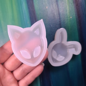 May include: Two clear silicone molds, one in the shape of a fox head and the other a bunny head. The molds are held against a teal and green background. The fox mold has pointed ears and a triangular nose. The bunny mold has long ears.