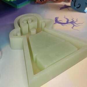 May include: A light green silicone mold with a unique shape, featuring a maze-like design and rectangular compartments. The mold is set against a backdrop of other colorful craft supplies, suggesting a creative project.