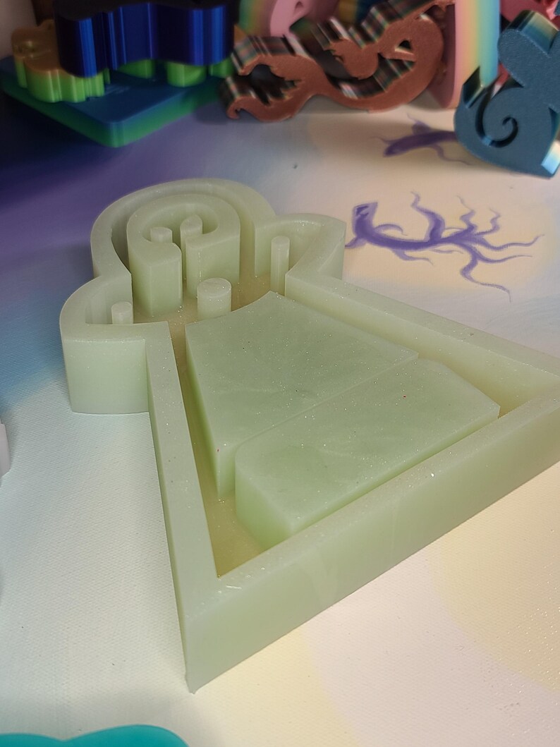 May include: A light green silicone mold with a unique shape, featuring a maze-like design and rectangular compartments. The mold is set against a backdrop of other colorful craft supplies, suggesting a creative project.