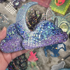 May include: A collection of glittery resin art pieces, including a cloud and crescent moon, in shades of blue, purple, and silver. Other items include alien head shapes and a butterfly. The pieces are likely handmade and suitable for crafts.
