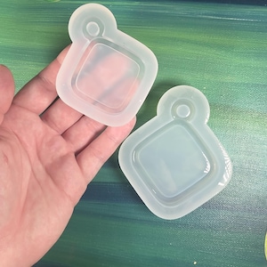 May include: Two clear, flexible silicone molds for crafting. Each mold has a square shape with rounded corners and a small loop at the top. The molds are held against a painted background of green and blue.