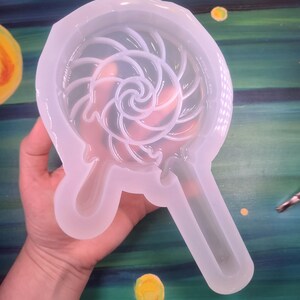 May include: A clear, flexible silicone mold in the shape of a lollipop. The mold features a spiral design within the lollipop shape and a handle. The mold is held in a hand against a colorful painted background.