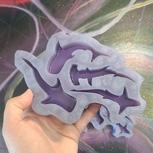 May include: A clear, irregular-shaped mold with a purple shark fin design. The mold is held in a hand, and the background is a colorful, abstract painting.