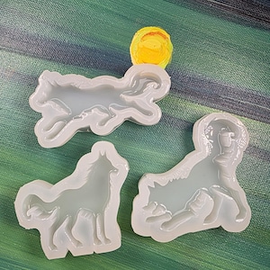 May include: Three clear silicone molds in the shape of wolves. One mold shows a wolf running, another shows a wolf standing, and the third shows a wolf lying down. The molds are on a green and blue painted background.