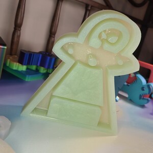 May include: A light green, translucent, UFO-shaped mold with a rectangular base and a rounded top. The mold has a textured surface and is filled with a clear, liquid-like substance. The design includes a stylized depiction of a flying saucer.