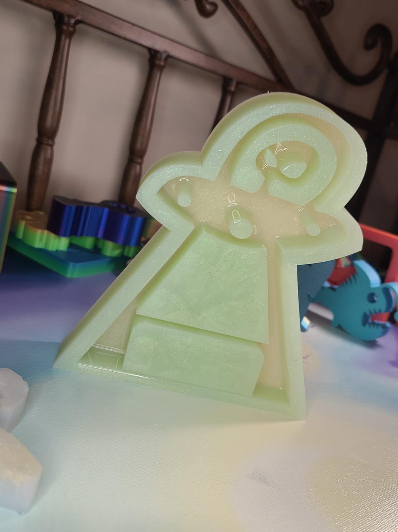 May include: A light green, translucent, UFO-shaped mold with a rectangular base and a rounded top. The mold has a textured surface and is filled with a clear, liquid-like substance. The design includes a stylized depiction of a flying saucer.
