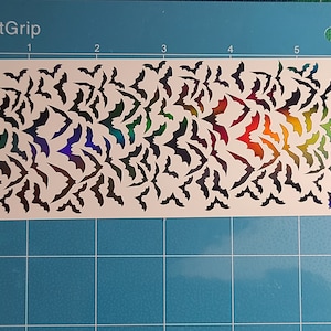 Peut inclure: Une feuille blanche avec un motif de chauves-souris d&eacute;coup&eacute;es en arc-en-ciel iridescent. La feuille est sur un fond de grille bleue.