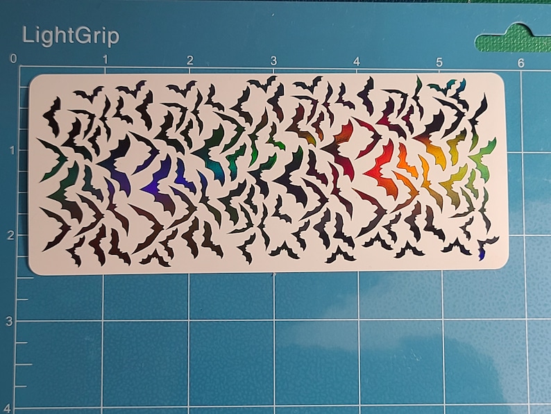 Peut inclure: Une feuille blanche avec un motif de chauves-souris d&eacute;coup&eacute;es en arc-en-ciel iridescent. La feuille est sur un fond de grille bleue.