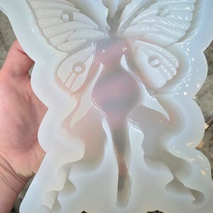 May include: A translucent, off-white silicone mold featuring a central figure with large, detailed butterfly wings. The design includes intricate segmented wing patterns and a smooth, elongated body shape, creating a hollow cavity for casting.