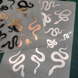 May include: Two sheets of stickers featuring gold and black snake designs. The stickers are on a gray background.