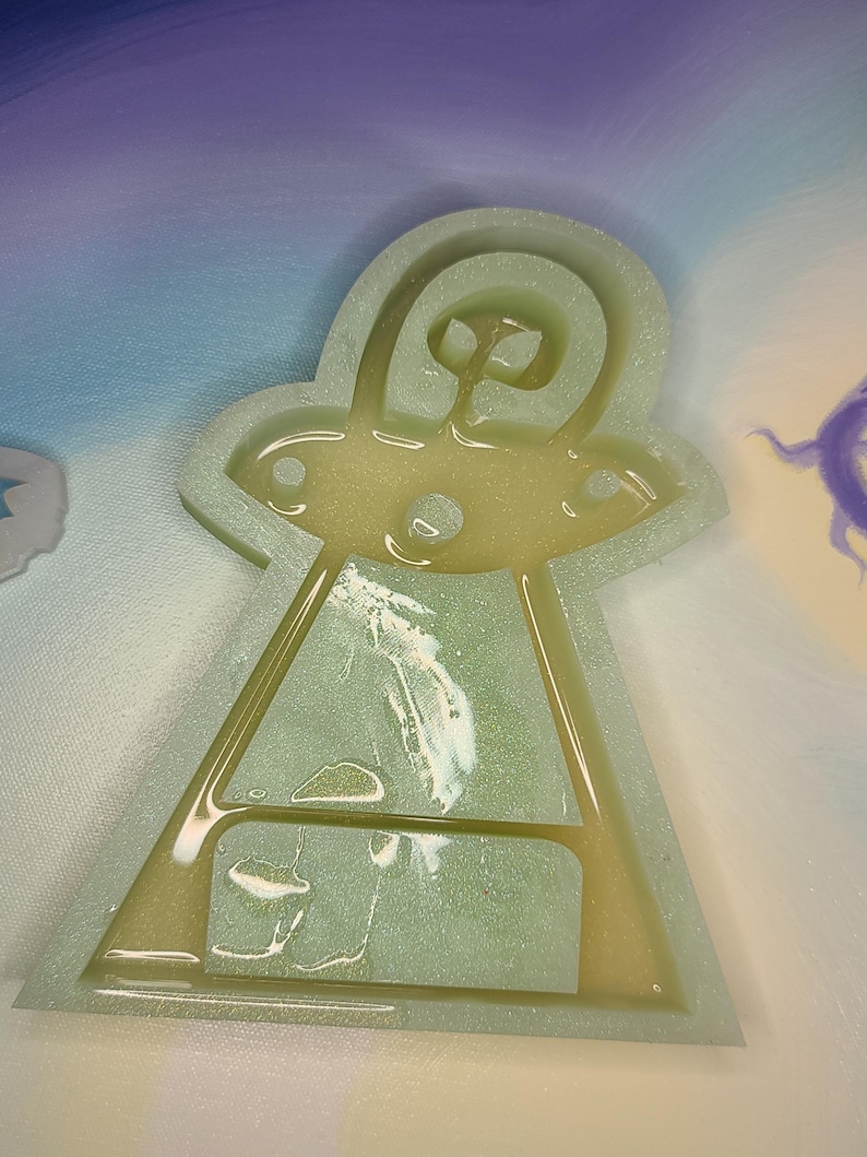 May include: A green and white glitter resin mold of a UFO. The mold is filled with resin and has a glittery finish.