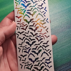 Peut inclure: Une feuille de papier autocollant blanc avec une finition arc-en-ciel iris&eacute;e. Le papier est rempli d'un motif de silhouettes de chauves-souris.