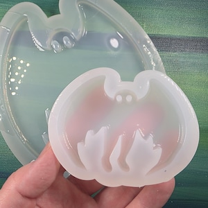 May include: A clear silicone mold in the shape of a bat, held in a hand. The bat mold is in front of a larger, similar mold. The background is a green and blue painted canvas.