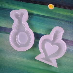 May include: Two white silicone molds, one shaped like a heart-shaped bottle and the other like a heart-shaped playing card suit. The molds are on a green and blue painted background.