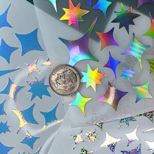 以下が含まれることがあります： ホログラフィックやグリッターの質感を含む、さまざまなサイズと仕上げの星型ステッカーのコレクション。白い台紙に配置されています。スケールの参考として、ステッカーの間にコインが置かれています。