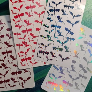 May include: Four sheets of stickers featuring various dragon designs. The stickers are on white cardstock and feature red, black, and iridescent holographic dragons.
