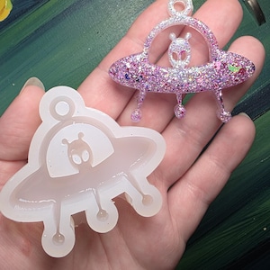 May include: A clear silicone mold and a finished glittery alien spaceship pendant. The pendant is pink and purple with an alien figure inside. The mold is in the shape of the spaceship and alien.