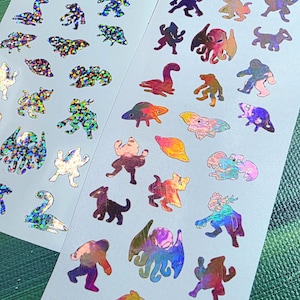 May include: Two rectangular sticker sheets featuring an array of small, holographic die-cut designs. The stickers depict various creatures, cryptids, and unidentified flying objects in shimmering, multi-colored metallic finishes on white backing.