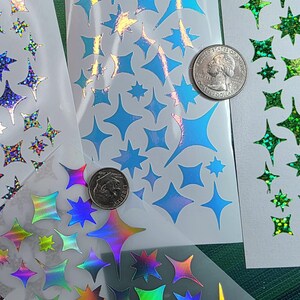 以下が含まれることがあります： 様々なサイズの星型ステッカーシート3枚。1枚は青いホログラムの星、1枚は緑のグリッターの星、もう1枚は虹色に輝く星がデザインされています。サイズ比較のためにアメリカの25セント硬貨が置かれています。