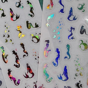 May include: Sheets of holographic mermaid stickers in various colors, including blue, purple, and gold. The stickers feature mermaid silhouettes and bubbles. The stickers are on a white background.