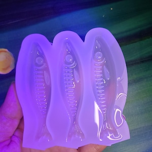 May include: A clear silicone mold featuring three fish-shaped cavities. The mold is held in a hand, with the fish designs visible. The background is a mix of green and blue paint.