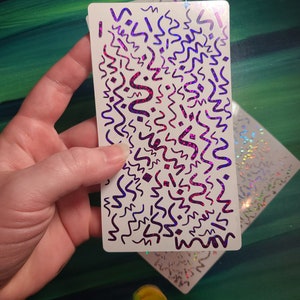 May include: A white sticker sheet with a holographic finish. The sticker sheet features a repeating pattern of purple and pink abstract shapes on a white background.