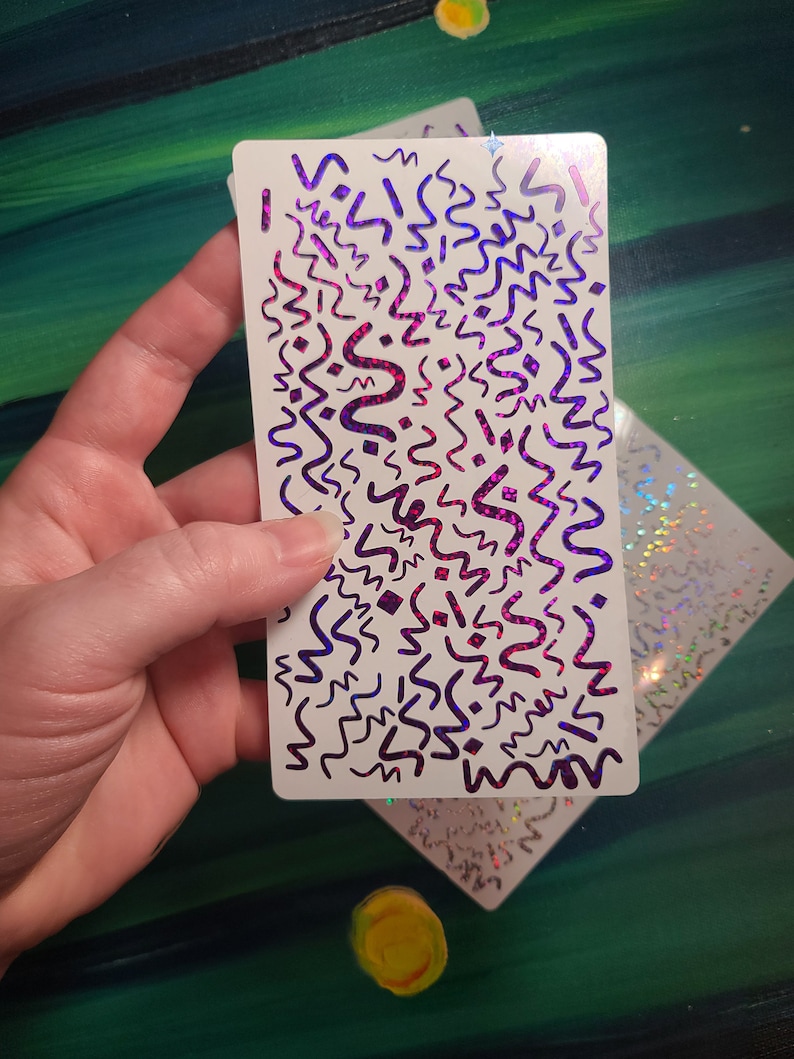 May include: A white sticker sheet with a holographic finish. The sticker sheet features a repeating pattern of purple and pink abstract shapes on a white background.