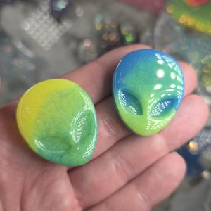 May include: Two alien-shaped resin pendants with a gradient of colors. One is yellow and green, the other blue and green. The pendants have detailed facial features and are likely for jewelry or crafting.
