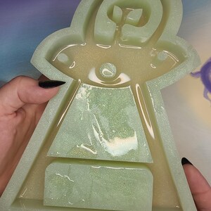 May include: A light green, glittery resin mold in the shape of a UFO. The mold has a detailed design with a central eye and a rectangular base. The edges are slightly rounded, and the overall shape is symmetrical.