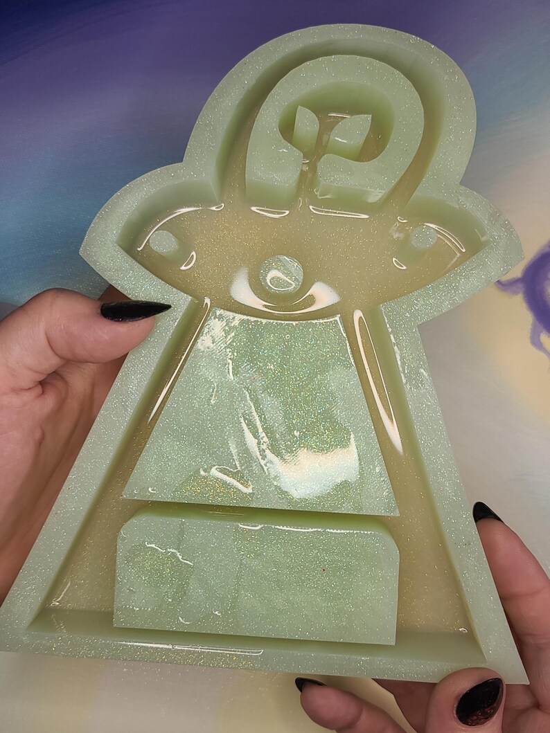 May include: A light green, glittery resin mold in the shape of a UFO. The mold has a detailed design with a central eye and a rectangular base. The edges are slightly rounded, and the overall shape is symmetrical.