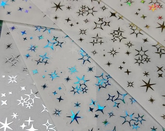 Puede incluir: Primer plano de varias tiras de pegatinas con forma de estrella en varios colores, incluyendo plata, negro e iridiscente. Las pegatinas están dispuestas sobre un fondo blanco, con una variedad de diseños de estrellas.