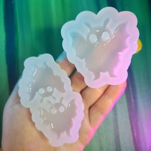 May include: Two translucent silicone bat-shaped molds. The molds have a rounded shape with bat wing details and two small eye details. The molds are held in a hand against a colorful background.