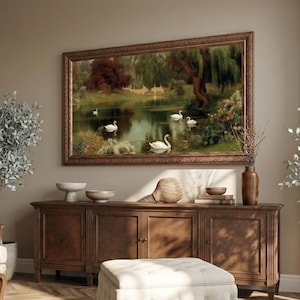 Pode incluir: Uma pintura de paisagem retangular numa moldura de madeira escura representando um lago com cisnes brancos, árvores verdes exuberantes e uma ponte de pedra. A obra está pendurada sobre um aparador de madeira comprido com quatro portas.