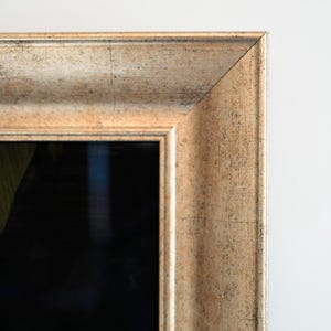 Pode incluir: Close-up de uma moldura dourada com um acabamento texturizado e desgastado. A moldura tem uma forma retangular clássica com uma borda chanfrada. O canto da moldura é visível contra um fundo branco, destacando os detalhes.