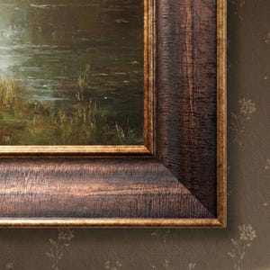 Op de afbeelding: Een ingelijst landschapsschilderij met een donkerbruine, getextureerde houten lijst. Het schilderij toont een waterpartij met groene en bruine vegetatie. De lijst heeft een gouden binnenrand.