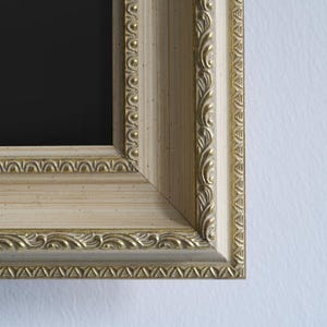Puede incluir: Primer plano de una esquina de un marco de fotos color crema con detalles dorados. El marco tiene un diseño detallado y ornamentado con un interior negro. El marco es de madera y tiene un acabado texturizado.