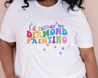 Può includere: Maglietta bianca con il testo "i'd rather be DIAMOND PAINTING" in colori arcobaleno. Il testo è in uno stile di font retrò. Ci sono piccole forme di diamanti color arcobaleno sparse intorno al testo.