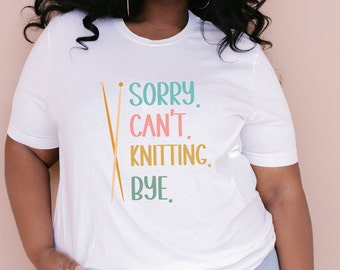 May include: White t-shirt with the text "Sorry. Can't. Knitting. Bye." in colorful lettering and a pair of crossed knitting needles.